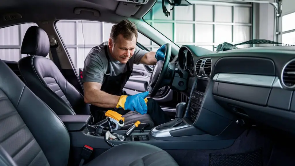 A meticulously detailed photograph of a car owner diligently cleaning the interior of his car. The owner is seated in the driver's seat, using various cleaning tools and kits by his side. The dashboard and controls are spotless, and the seats are immaculate, showcasing the owner's dedication to maintaining the pristine condition of his vehicle. The background features a spacious garage with ample natural light, highlighting the car's gleaming exterior.