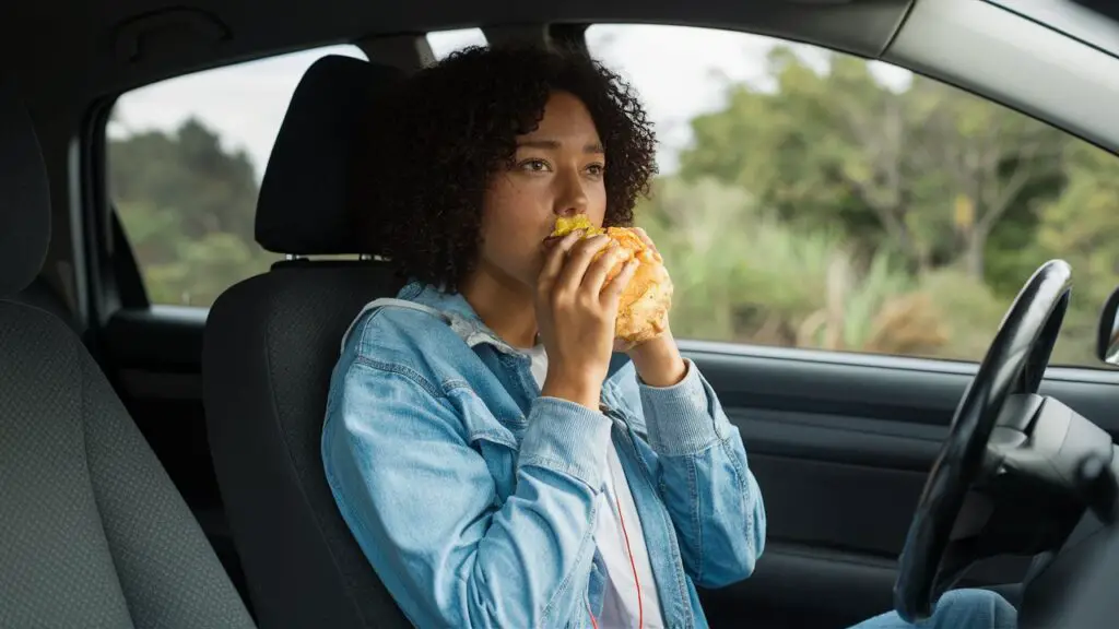 A lady on wig wearing blue jacket eating burger inside a car